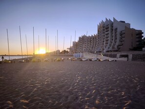 A beach with a row of white beach chairs and umbrellas, a modern building with balconies, and the sun setting in the background. - Le Castel · La Baule · 6 guests · near the beach (La Baule-Escoublac)