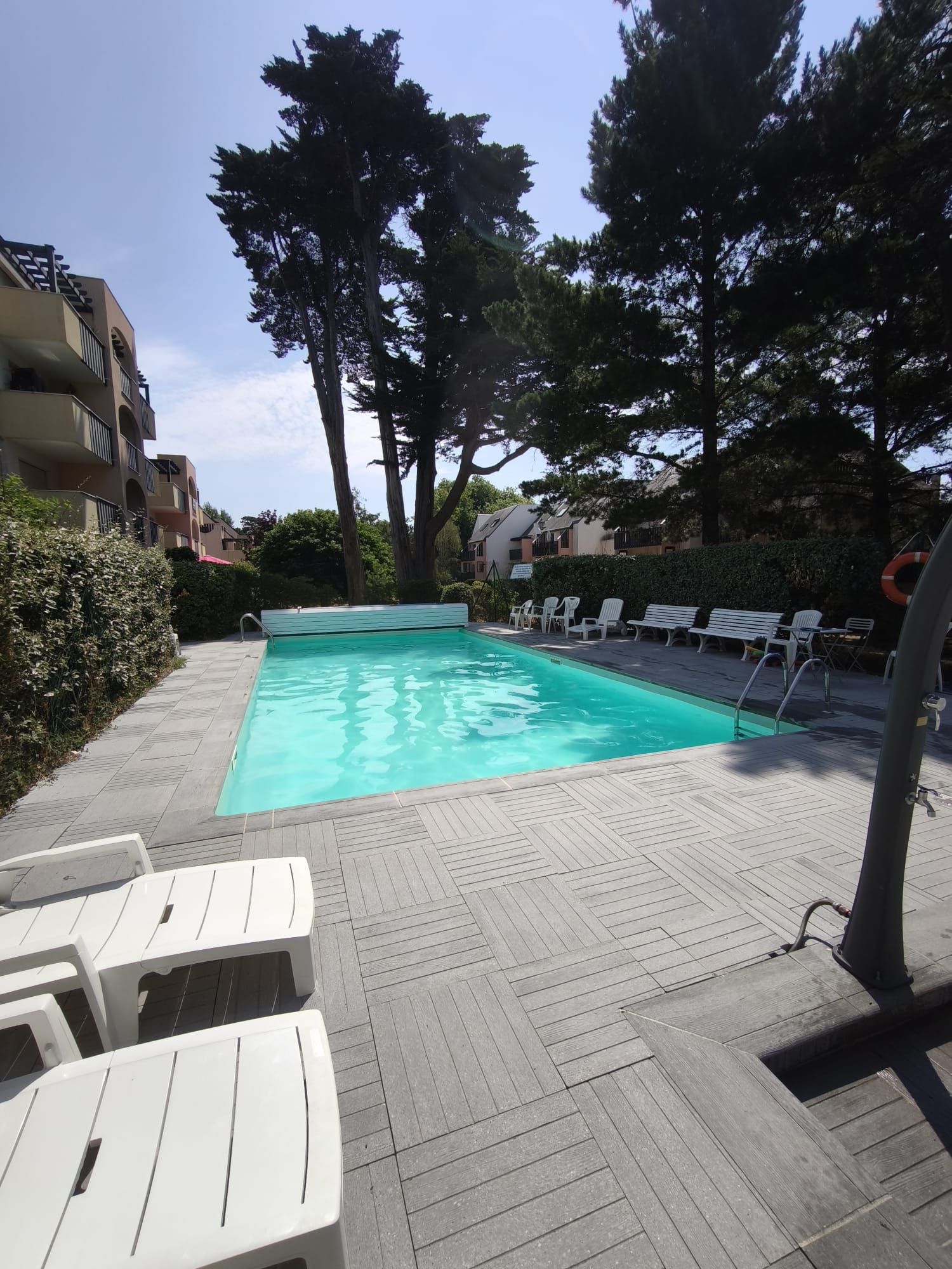 Une piscine entourée d’arbres et d’immeubles, avec des chaises longues et un cercle de sauvetage à proximité.