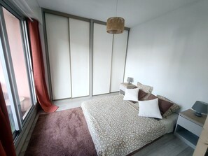 Ein Schlafzimmer mit Bett, Nachttischen, einer Pendelleuchte und Blick durch ein Fenster mit Vorhängen.