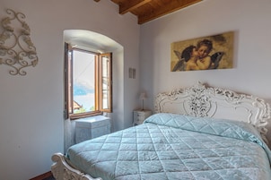 A bedroom with a bed, a window with a view, a nightstand, and a painting on the wall.