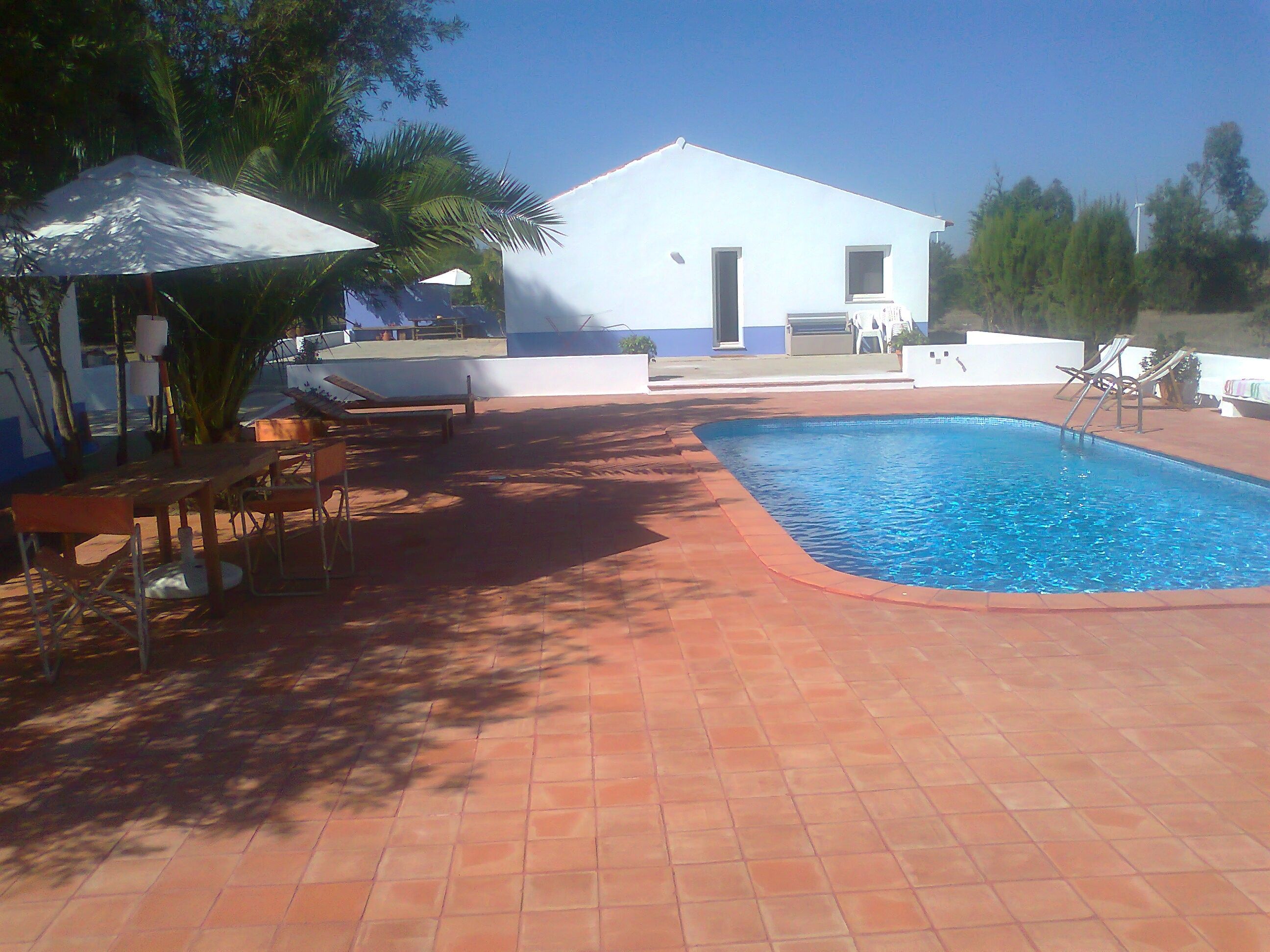 A swimming pool with a tiled deck, a white building with a door and windows, and outdoor furniture including chairs and a table.