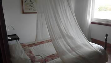 Ein Zimmer mit einem Bett, das mit einem transparenten weißen Moscheevorhang verziert ist, einem kleinen Nachttisch mit Lampe und einem gerahmten Kunstwerk an der Wand.