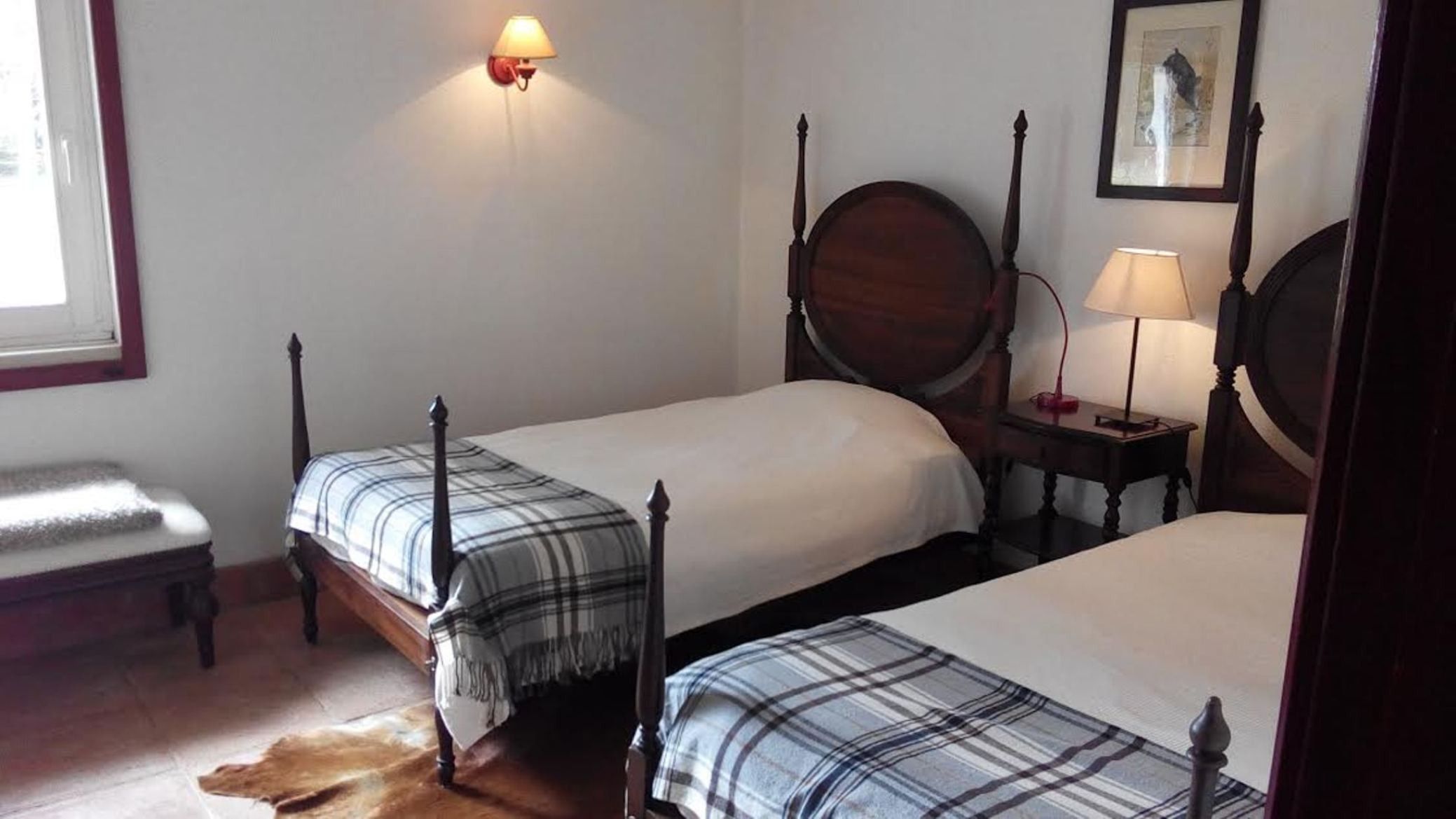 A room with two beds, a bench, a lamp, and a framed picture on the wall.