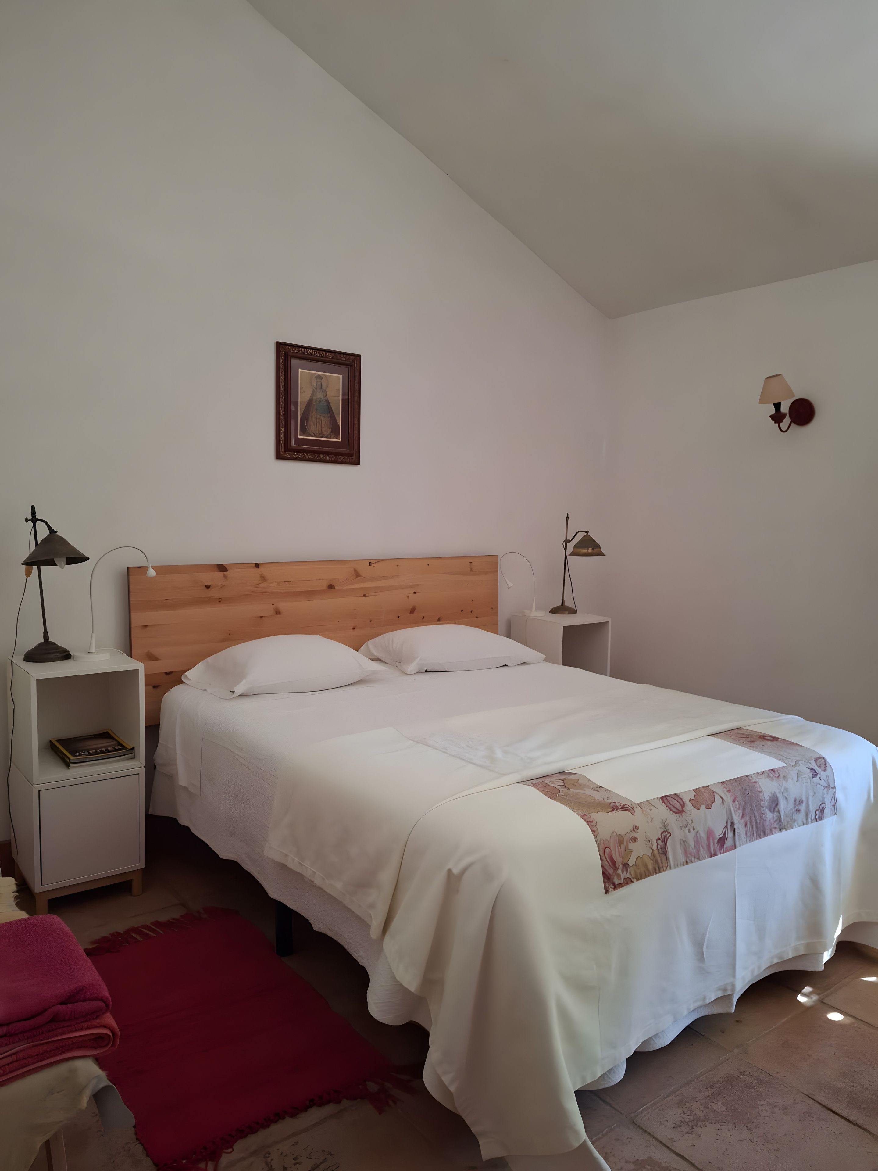 A bedroom with a wooden headboard, white bedding, a red throw blanket, and a framed picture on the wall.