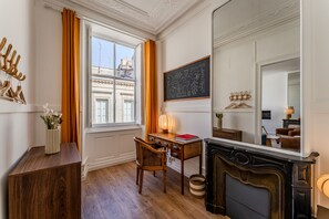 Quarto com escrivaninha de madeira, cadeira e lareira. Há uma janela com cortinas, um espelho e um cabideiro na parede.