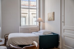 A bedroom with a bed, a bedside table, a lamp, and a view of buildings outside the window. - Triangle d'Or — Elegance & King Size bed (Bordeaux)