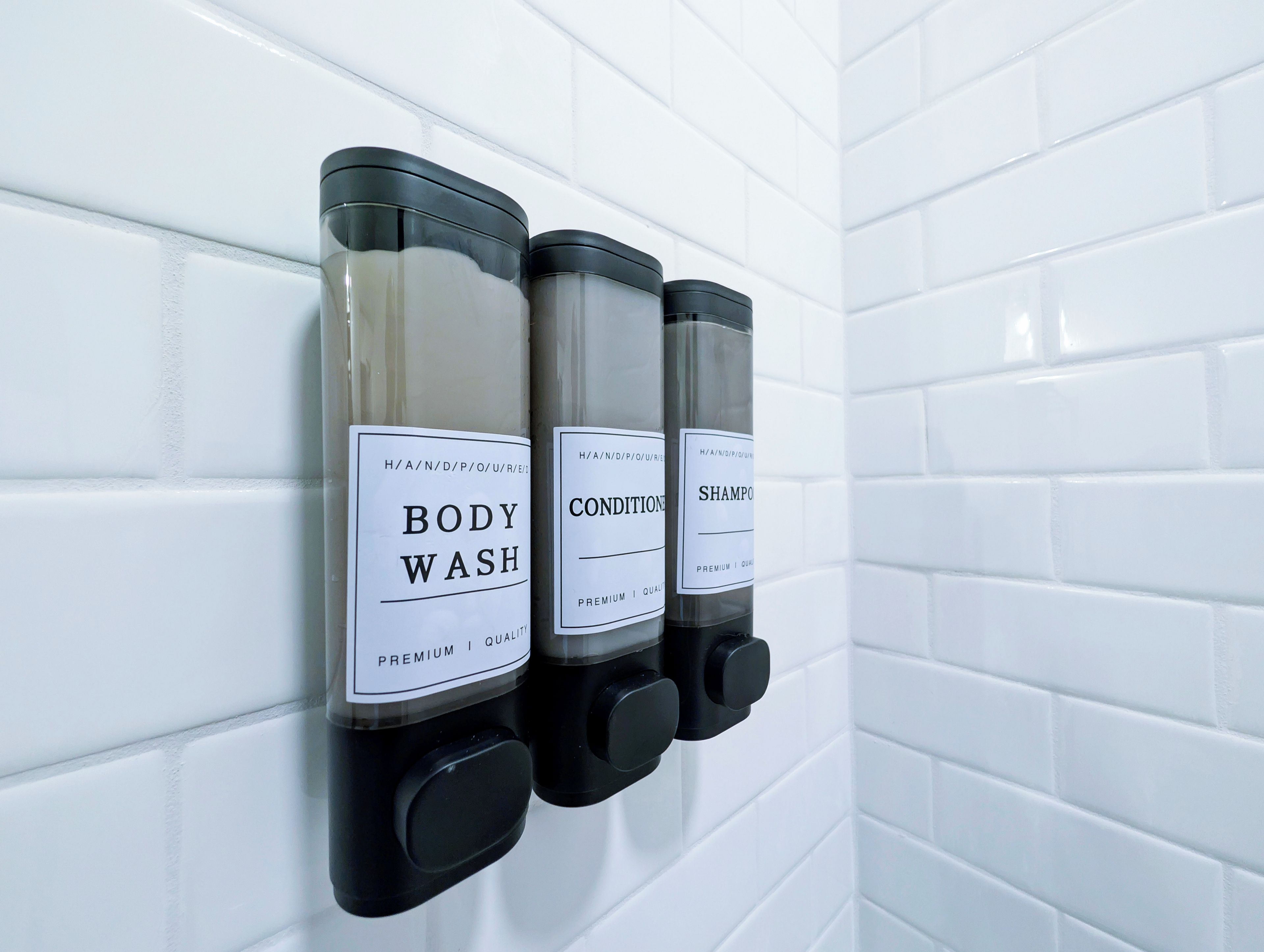 A wall-mounted dispenser with three bottles labeled 'BODY WASH,' 'CONDITIONER,' and 'SHAMPOO.'