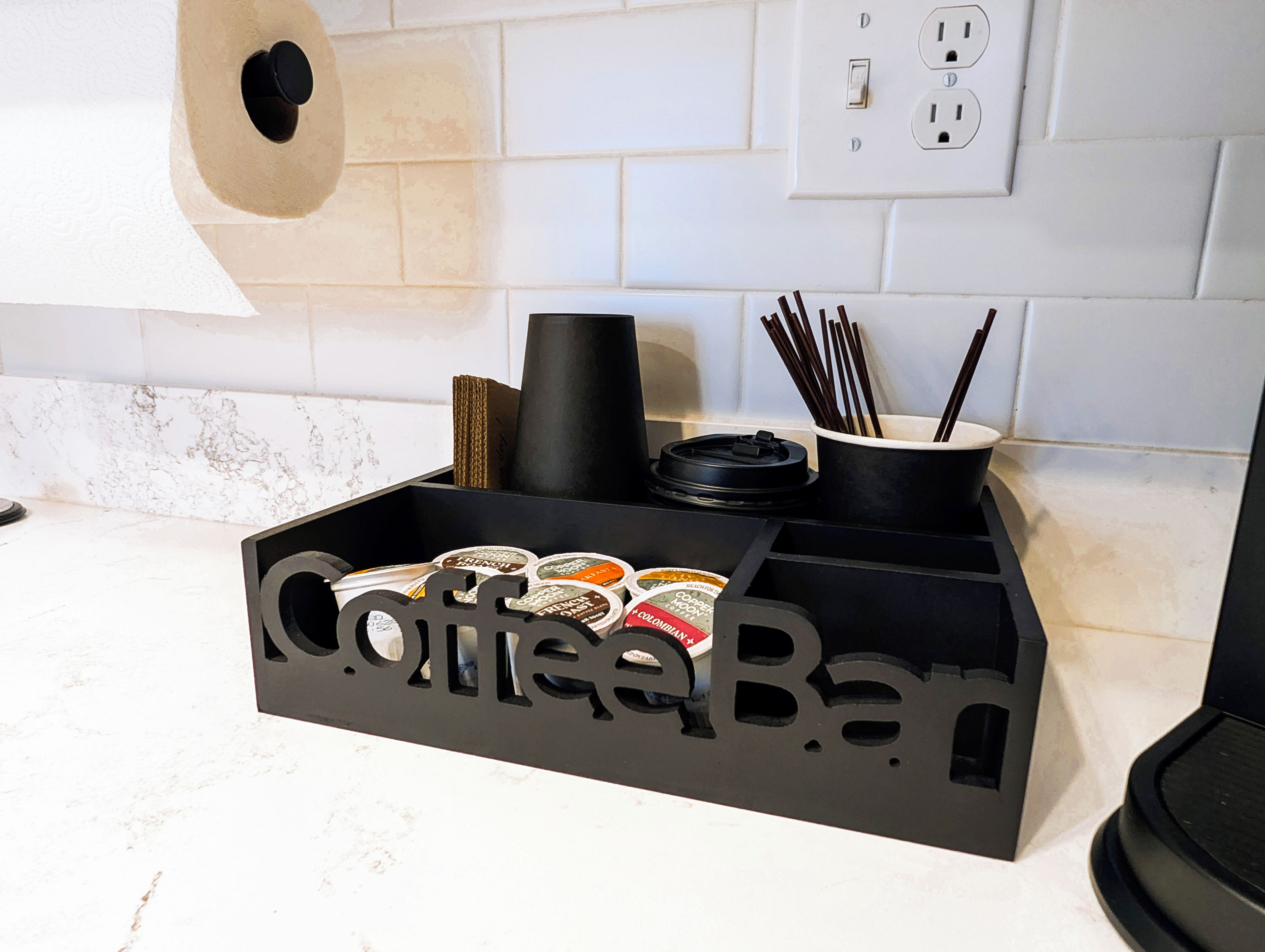 A kitchen counter with a black coffee bar containing coffee pods and a cup holder with chopsticks.