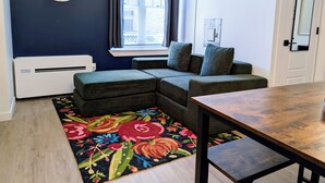 Un salon moderne avec un canapé d’angle, une table en bois, un tapis coloré, une horloge murale et une fenêtre avec des rideaux.