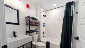 Une salle de bain moderne avec un lavabo, des toilettes et une baignoire, tous de couleur blanche. On y trouve une étagère avec des serviettes et des articles de toilette.