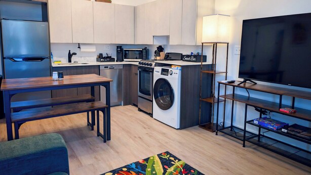 Une cuisine moderne équipée d’un four encastré, d’un four à micro-ondes et d’un lave-vaisselle, un coin repas avec une table en bois et un salon doté d’un téléviseur à écran plat.