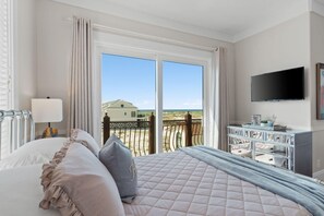 Quarto de hotel com uma cama grande, televisão na parede e vista para a praia pelas janelas.