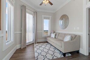 A room with a beige sofa, a patterned rug, a ceiling fan, and a view of the beach through a window. - Luxury Soundfront Home in Gated Community with Water Views, Pool & Fire Pit (Navarre Beach)