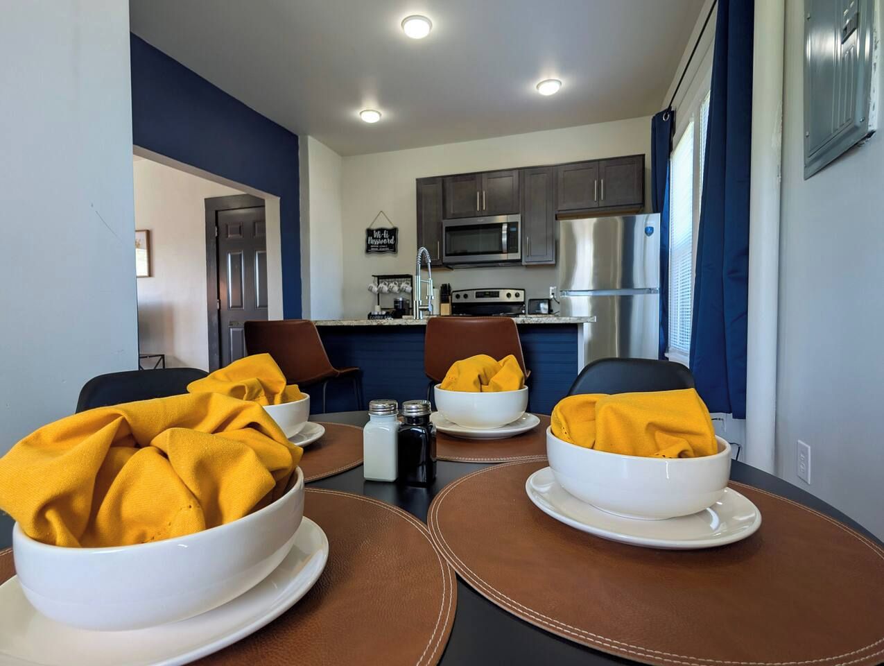Un comedor con una mesa dispuesta para cuatro personas, con cuencos blancos y servilletas amarillas.