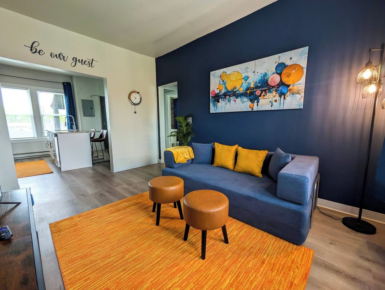 Una sala moderna con un sofá azul, dos otomanes marrones, un cojín amarillo y una pared con un letrero que dice 'be our guest'.