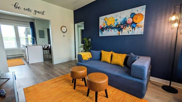 Una sala moderna con un sofá azul, dos otomanes marrones, un cojín amarillo y una pared con un letrero que dice 'be our guest'.