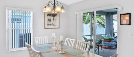 Un coin repas avec une table dressée pour quatre personnes, entourée de chaises blanches, d’un lustre et d’un tableau encadré accroché au mur.