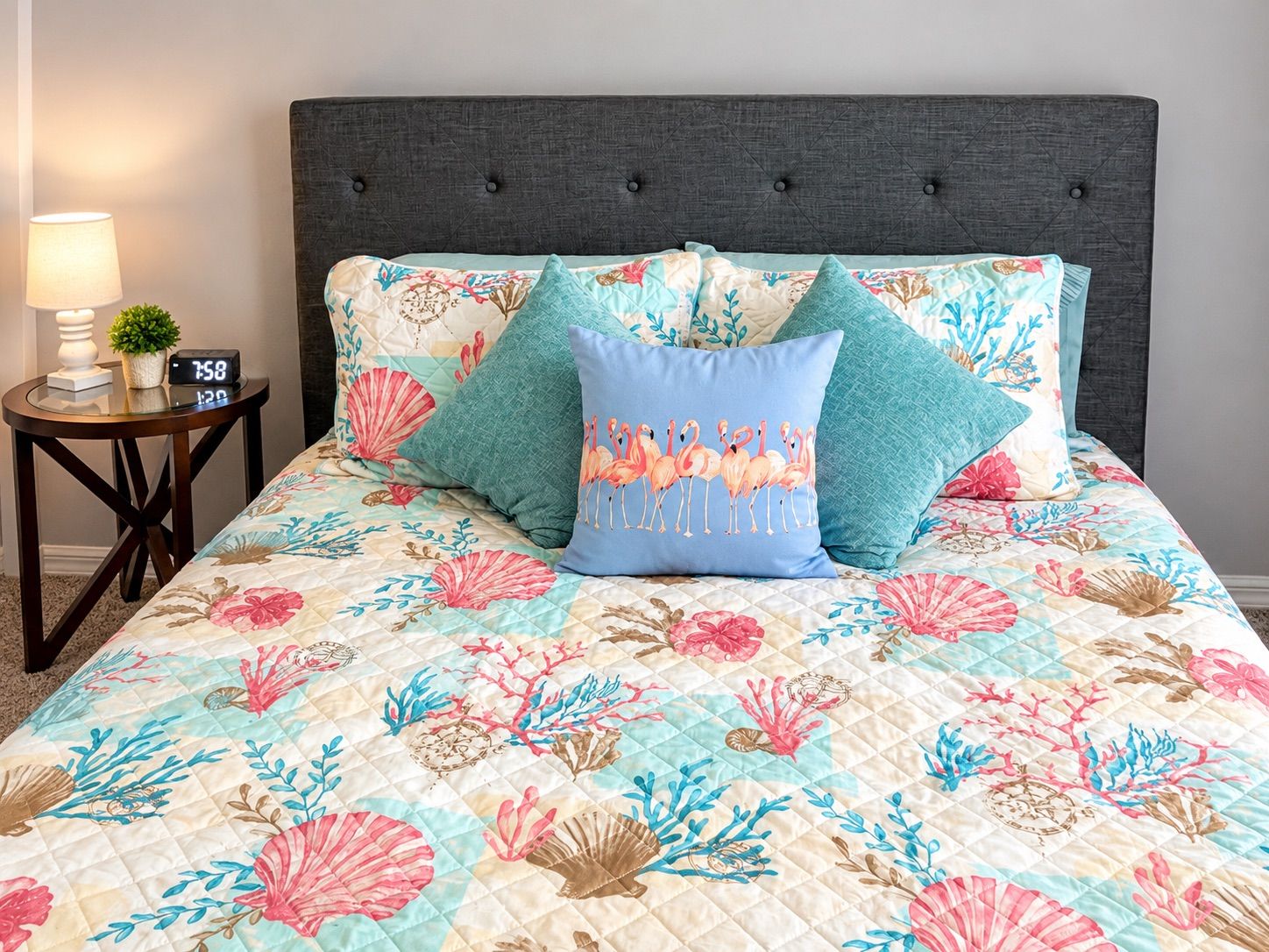 Een bed met een quilt met bloemenprint, een grijs gestoffeerd hoofdbord en een nachtkastje met een lamp en een klok.