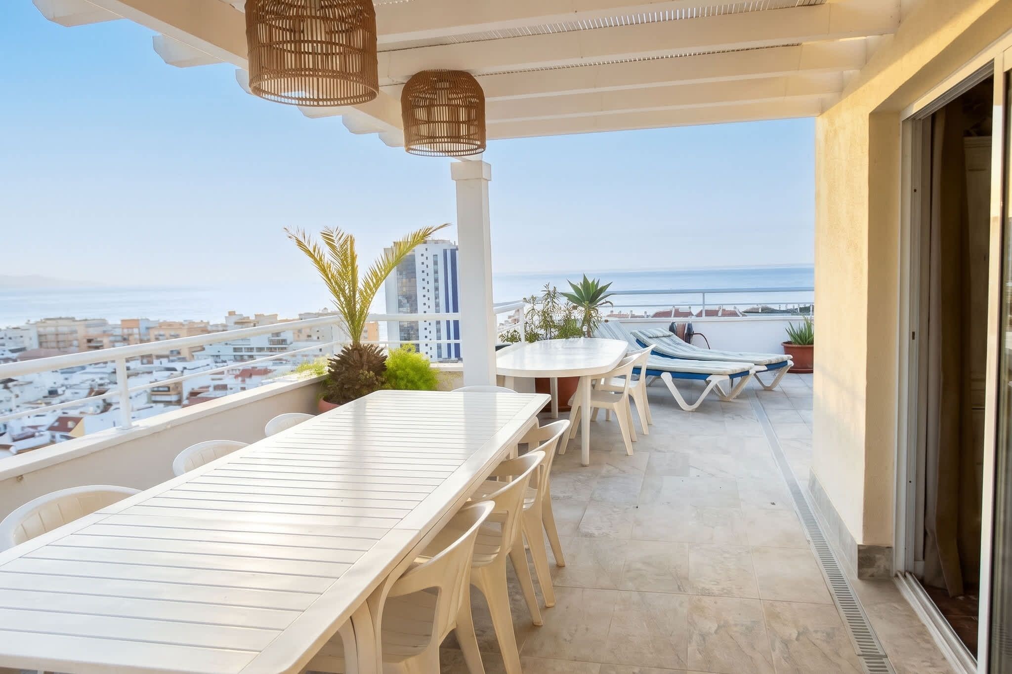 A balcony with a table and chairs, a hanging light fixture, and a view of the sea and buildings.