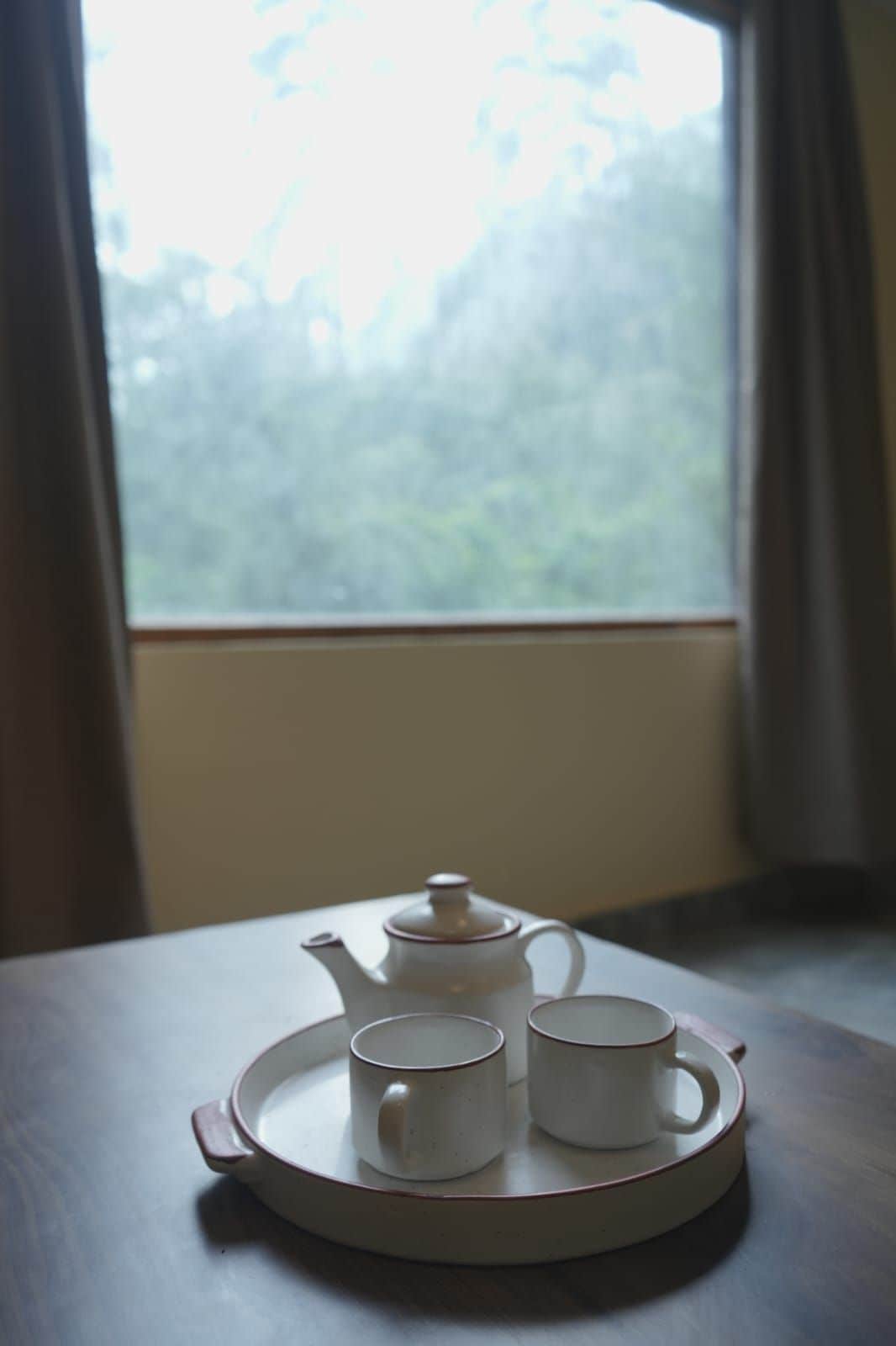 A white teapot and two white cups on a tray on a table by a window with curtains.