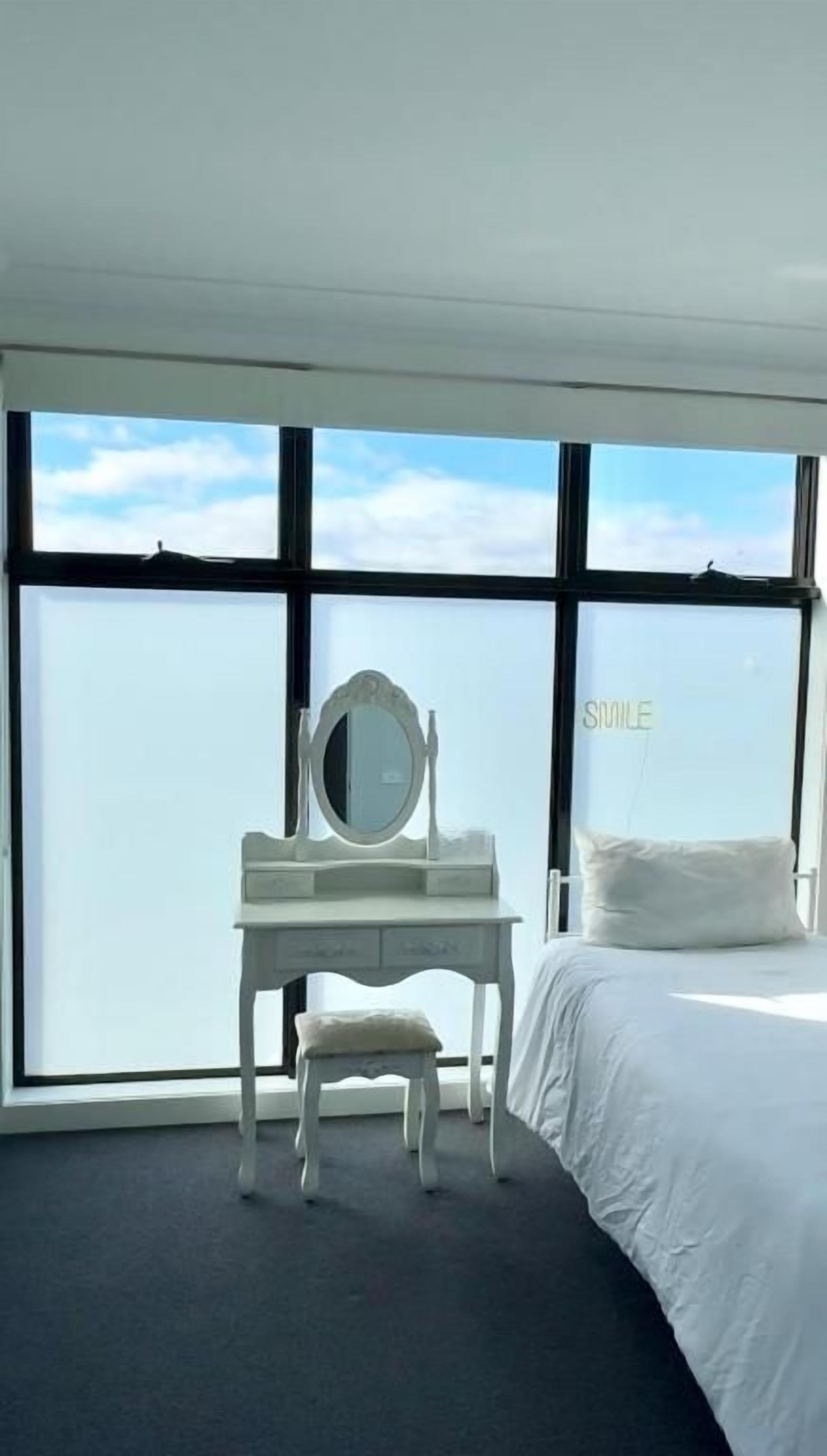 A bedroom with a bed, a white desk with a mirror, and a view through large windows.