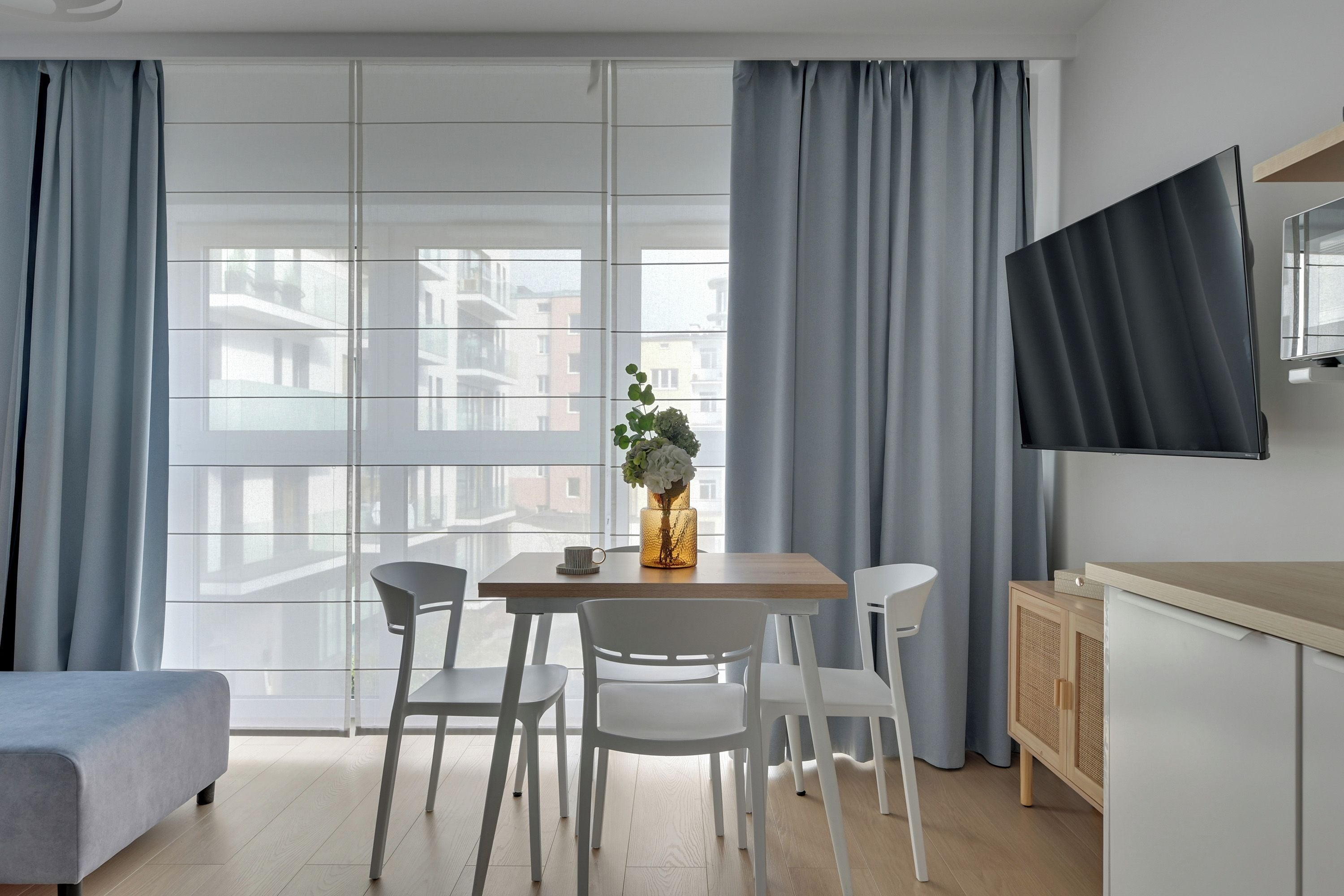A modern living room with a dining area, large windows, and a flat-screen TV mounted on the wall.