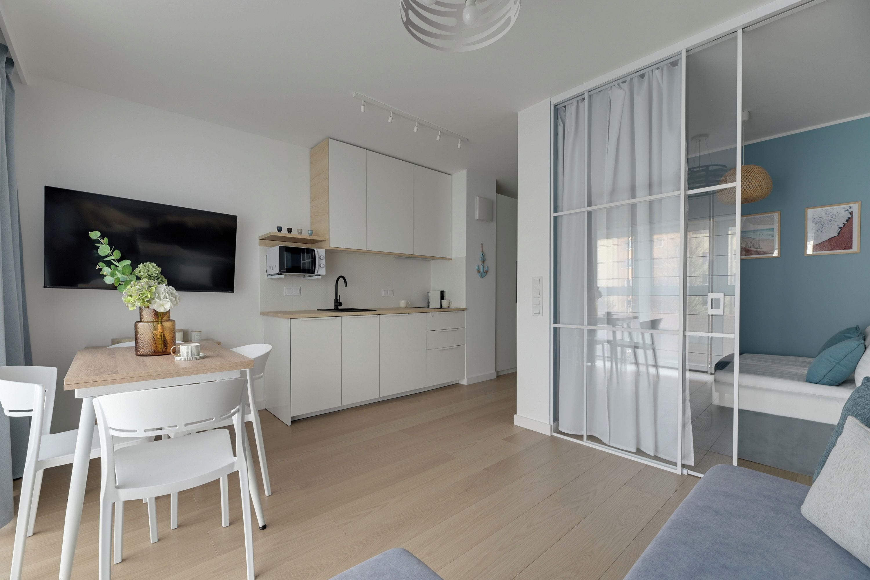 A modern kitchen with white cabinets, a dining area with a wooden table and white chairs, and a living area with a sofa and a glass partition.