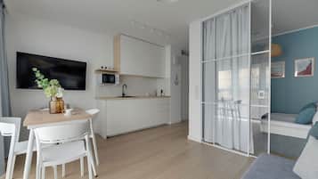 A modern kitchen with white cabinets, a dining area with a wooden table and white chairs, and a living area with a sofa and a glass partition.