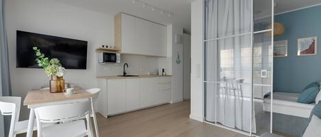 Una cocina moderna con armarios blancos, un comedor con mesa de madera y sillas blancas, y una sala de estar con sofá y una pared divisoria de vidrio.