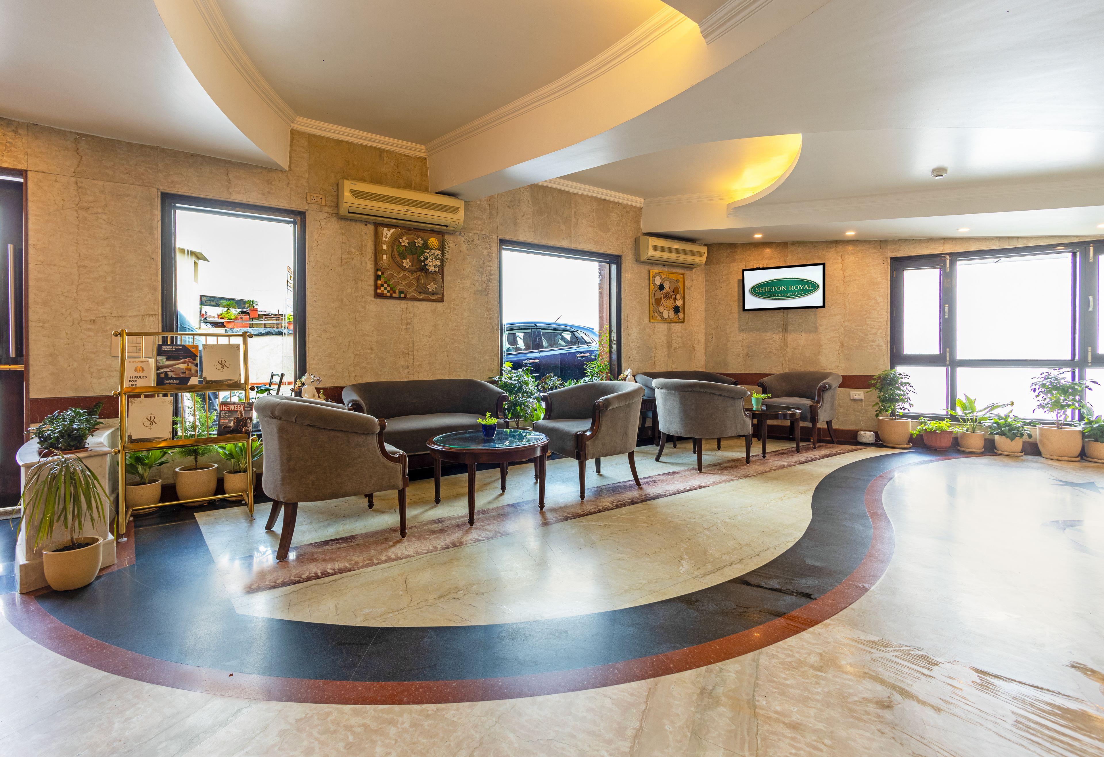 Vestíbulo de hotel con piso de mármol curvo, zona de estar con sofás y sillones, plantas en macetas y un ventanal con vistas al exterior.