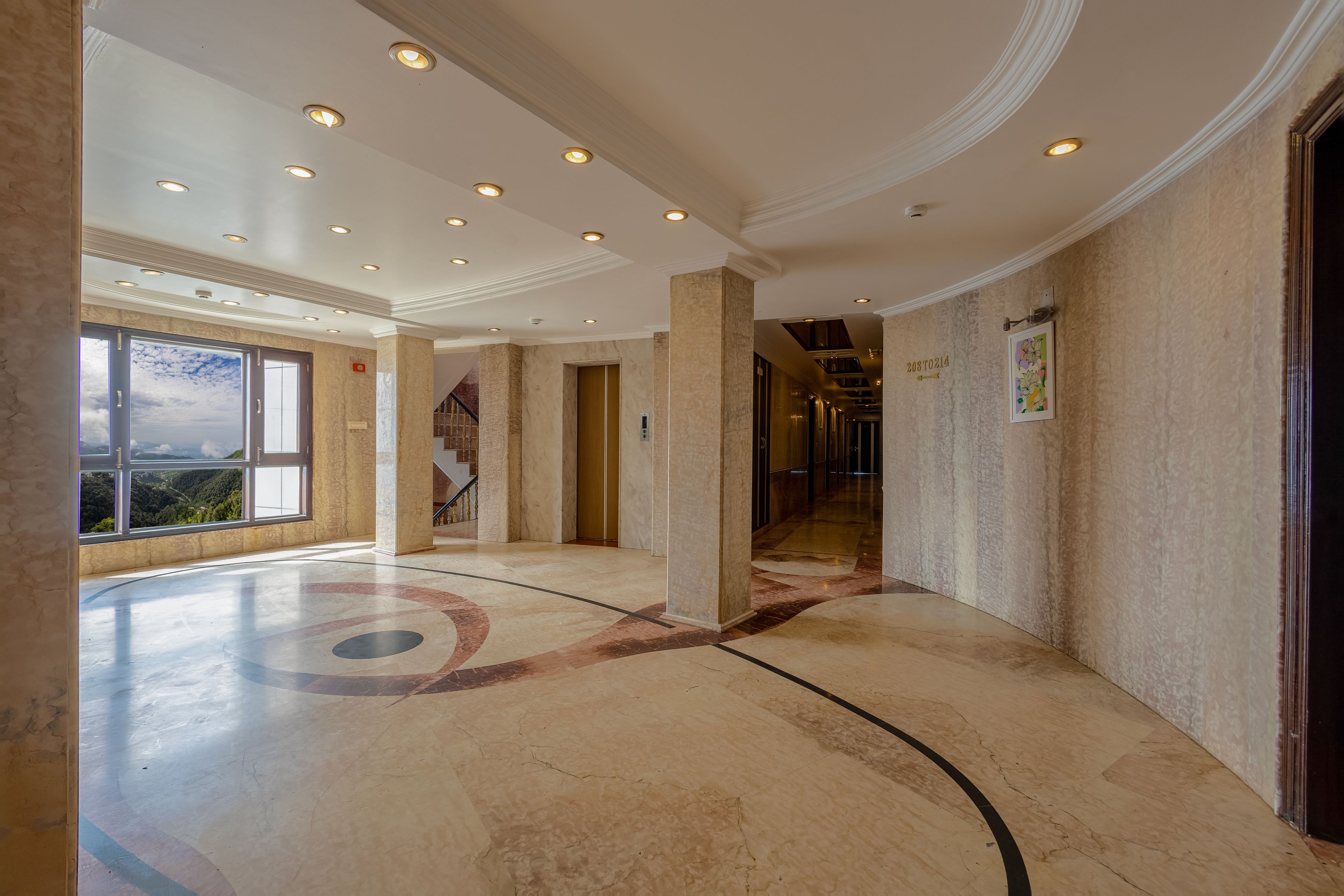 A spacious hallway with a marble floor, a large window offering a view of the outdoors, and a door with a number plaque.