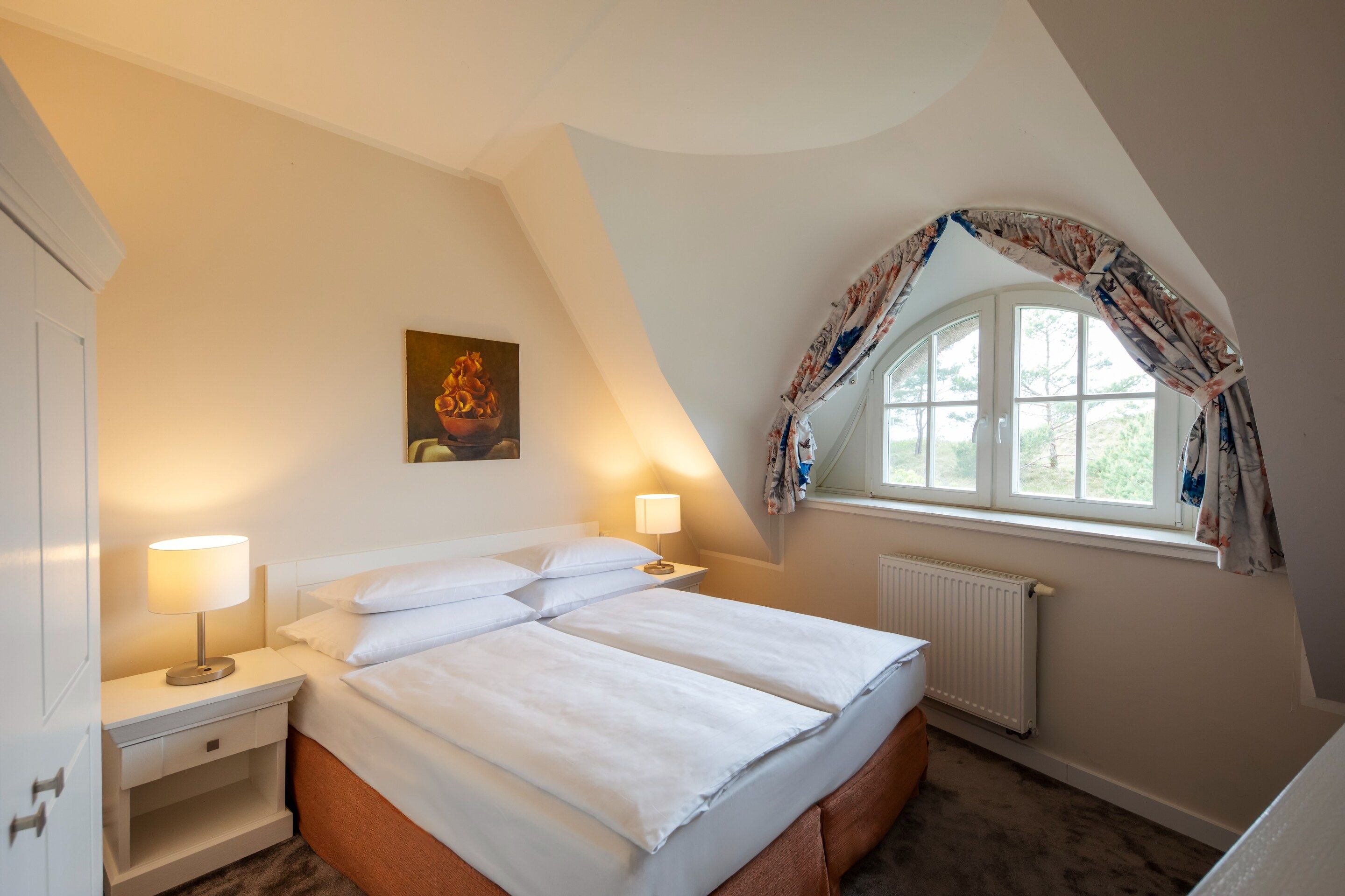 Una camera da letto con un letto, due comodini, un tavolo da notte, un termosifone, una finestra con tende e un quadro appeso al muro.