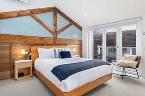 A bedroom with a wooden headboard, a bed with white bedding and a blue throw, a bedside table with a lamp, and a chair by the window. - Brick & Beam - Heritage Warehouse Richmond (Richmond)