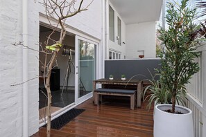 Een houten terras met een bank en potplanten, een witte muur en een schuifdeur met glas.