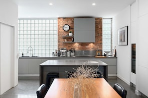 A modern kitchen with a brick wall, a clock, and a window with blinds. - Brick & Beam - Heritage Warehouse Richmond (Richmond)