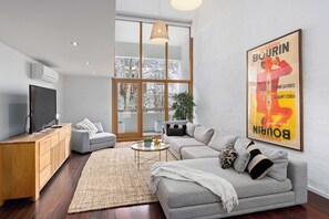A modern living room with a grey sofa, a television, and a large window. - Brick & Beam - Heritage Warehouse Richmond (Richmond)