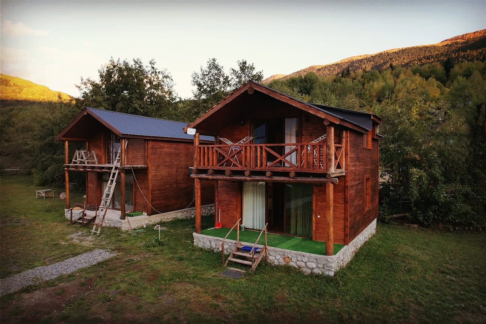 Two wooden cabins with balconies, set in a forested area with a mountain backdrop.