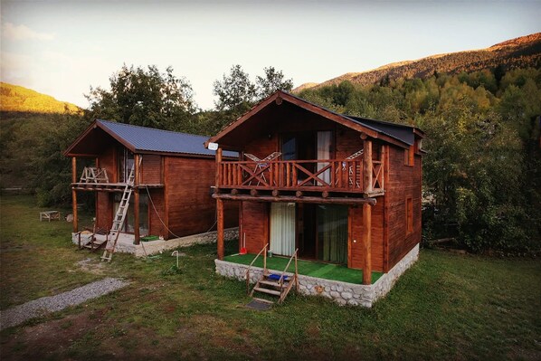 Two wooden cabins with balconies, set in a forested area with a mountain backdrop.