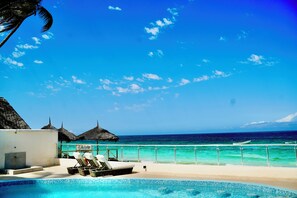 Un espace aménagé au bord de la piscine, avec des chaises longues, une structure à toit de chaume et une vue sur l’océan.