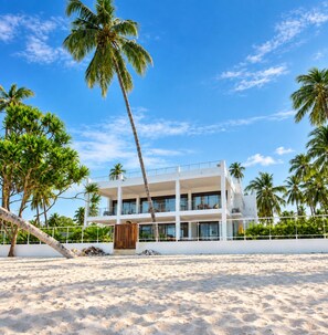 A beachfront hotel with multiple floors, large windows, and a palm tree in the foreground. - Exclusive 3-Bedroom Beachfront Villa with Private Pool in Zanzibar (Zanzibar)
