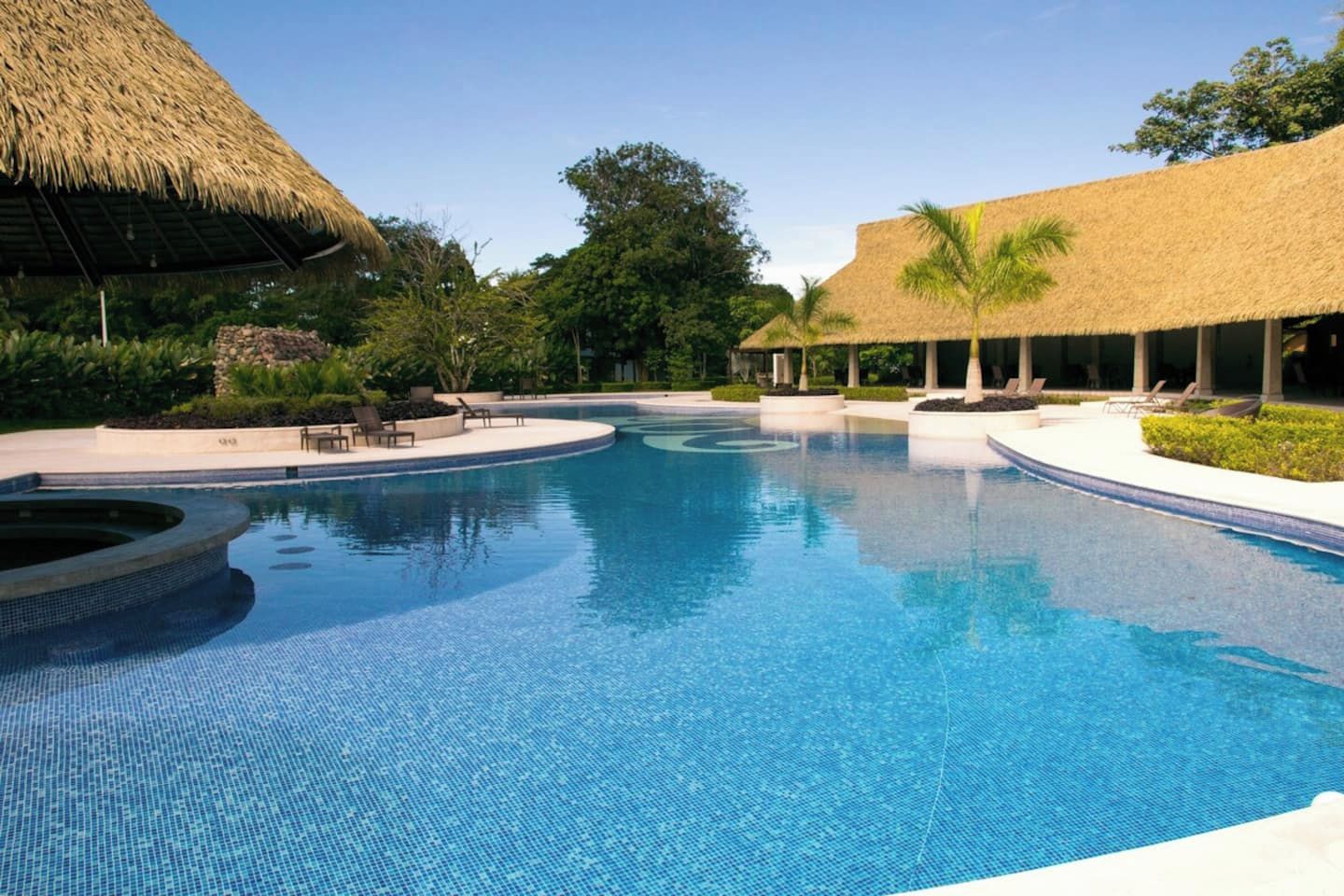 Ein Swimmingpool mit einer strohgedeckten Struktur an einer Seite, umgeben von Liegestühlen und viel Grün.