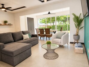 A modern living room with a grey sofa, a white armchair, wicker chairs, a glass coffee table, and a view of the outdoors through large windows. - Halani Beach House - Ciudad del Mar - King bed (Jaco, Ciudad del Mar)