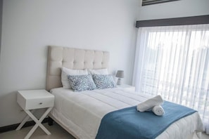 A hotel room with a bed, a nightstand, a lamp, and a window with curtains. - Halani Beach House - Ciudad del Mar - King bed (Jaco, Ciudad del Mar)