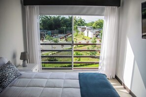 Ein Schlafzimmer mit einem großen Bett, einem Nachttisch mit Lampe und Blick auf eine angelegte Grünfläche mit Häusern.