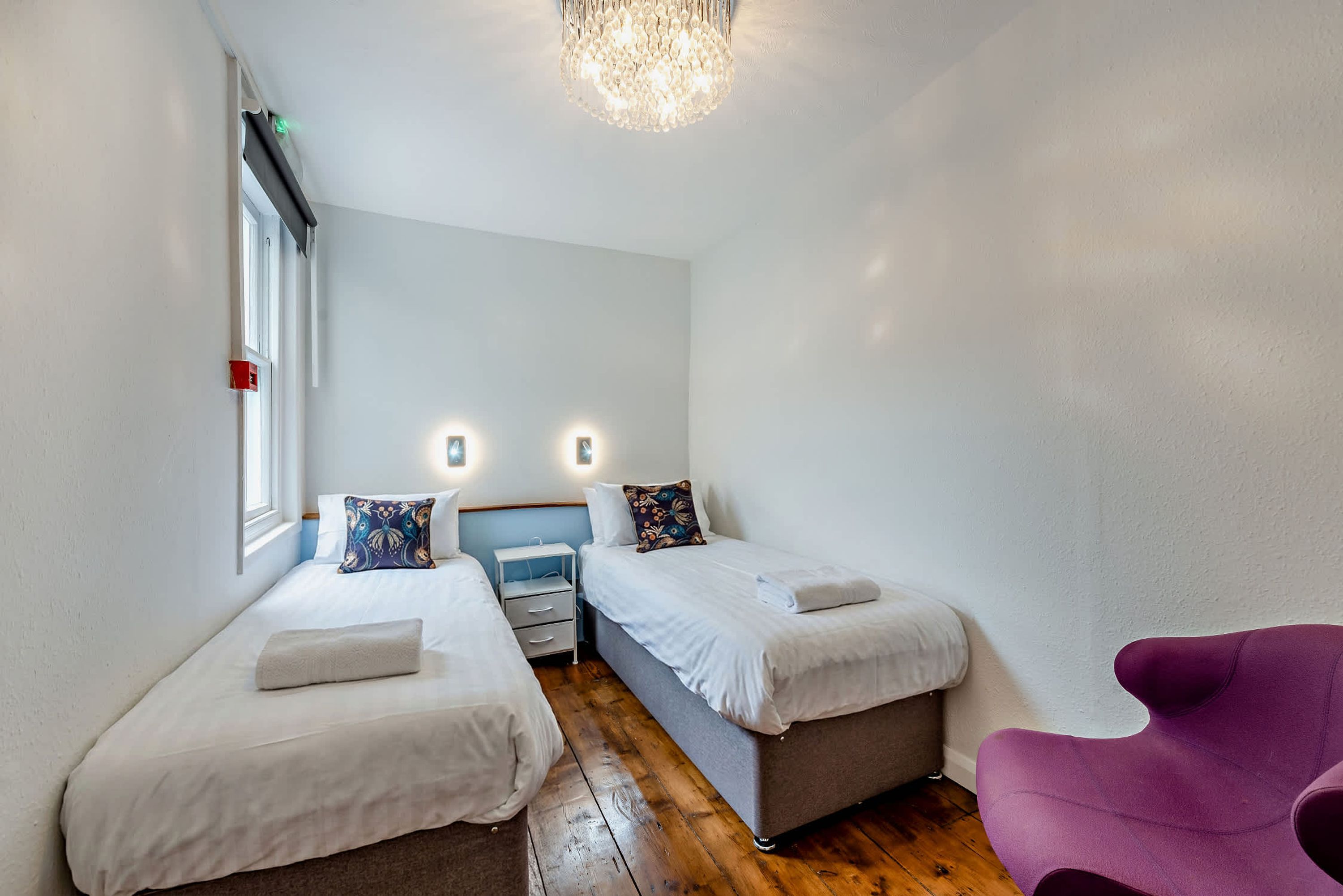 Een kleine, goed verlichte kamer met twee bedden, een nachtkastje, een stoel en een kroonluchter.