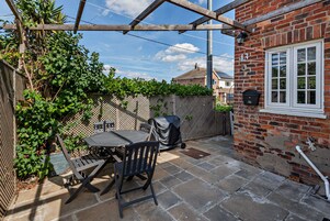 A patio with a table and chairs, a covered grill, and a brick wall with a window.