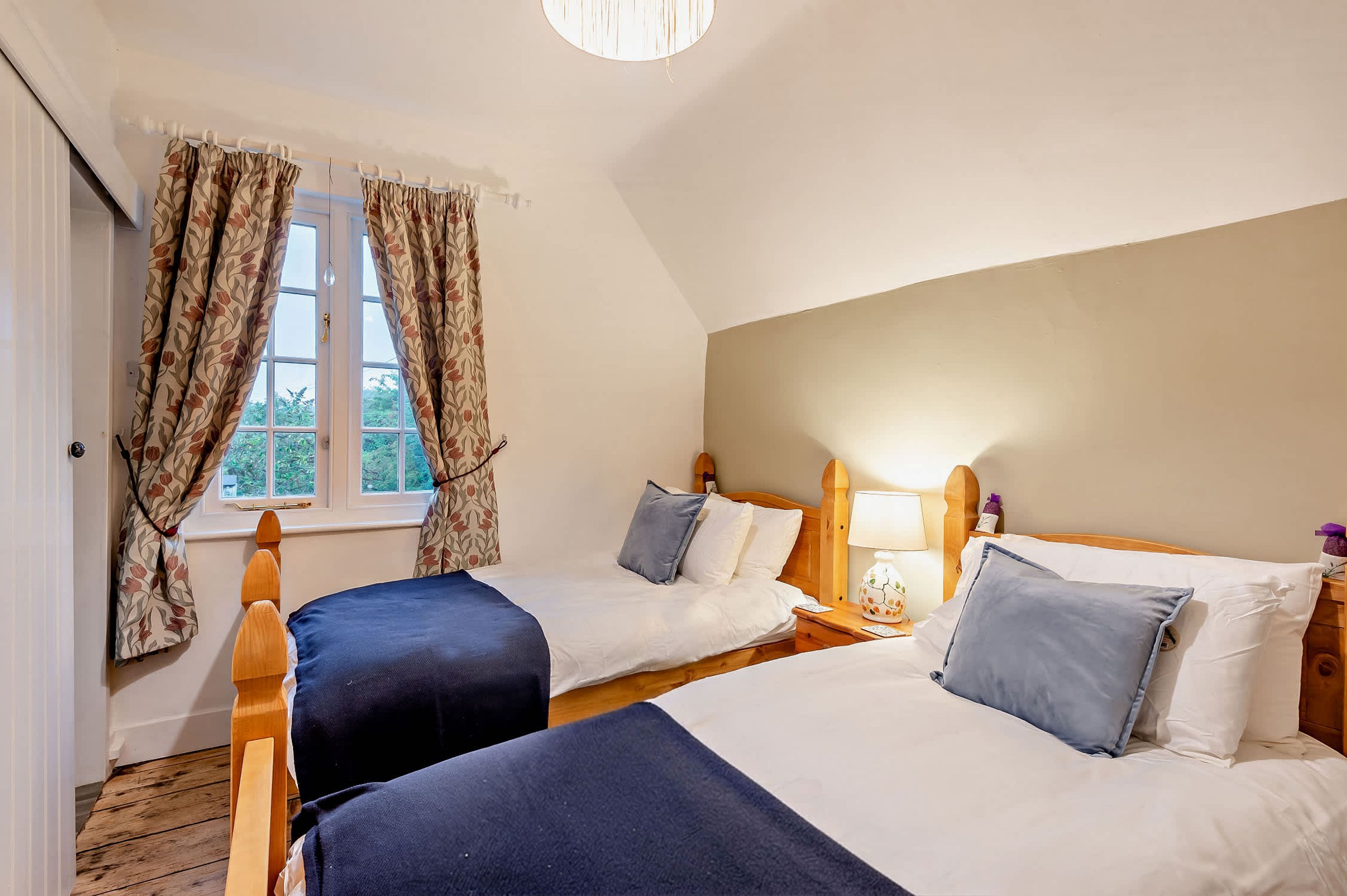 Une chambre avec deux lits, une fenêtre avec des rideaux, une tête de lit en bois et une table de chevet avec une lampe.
