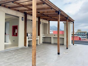 Un patio exterior con una pérgola de madera, una parrilla y vistas a la ciudad y montañas al fondo.