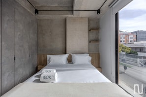 Ein modernes Schlafzimmer mit einem großen Bett, zwei Kissen, einem Nachttisch und Blick auf die Stadt durch ein Fenster.