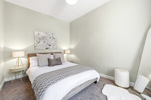 A bedroom with a bed, two bedside tables, a nightstand, a mirror, and a wall-mounted abstract painting.